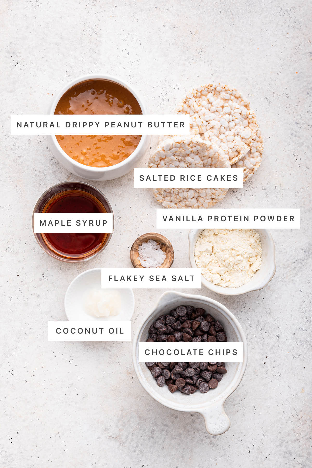 Ingredients measured out to make No Bake Peanut Butter Crunch Bars: natural drippy peanut butter, salted rice cakes, maple syrup, flakey sea salt, vanilla protein powder, coconut oil and chocolate chips.