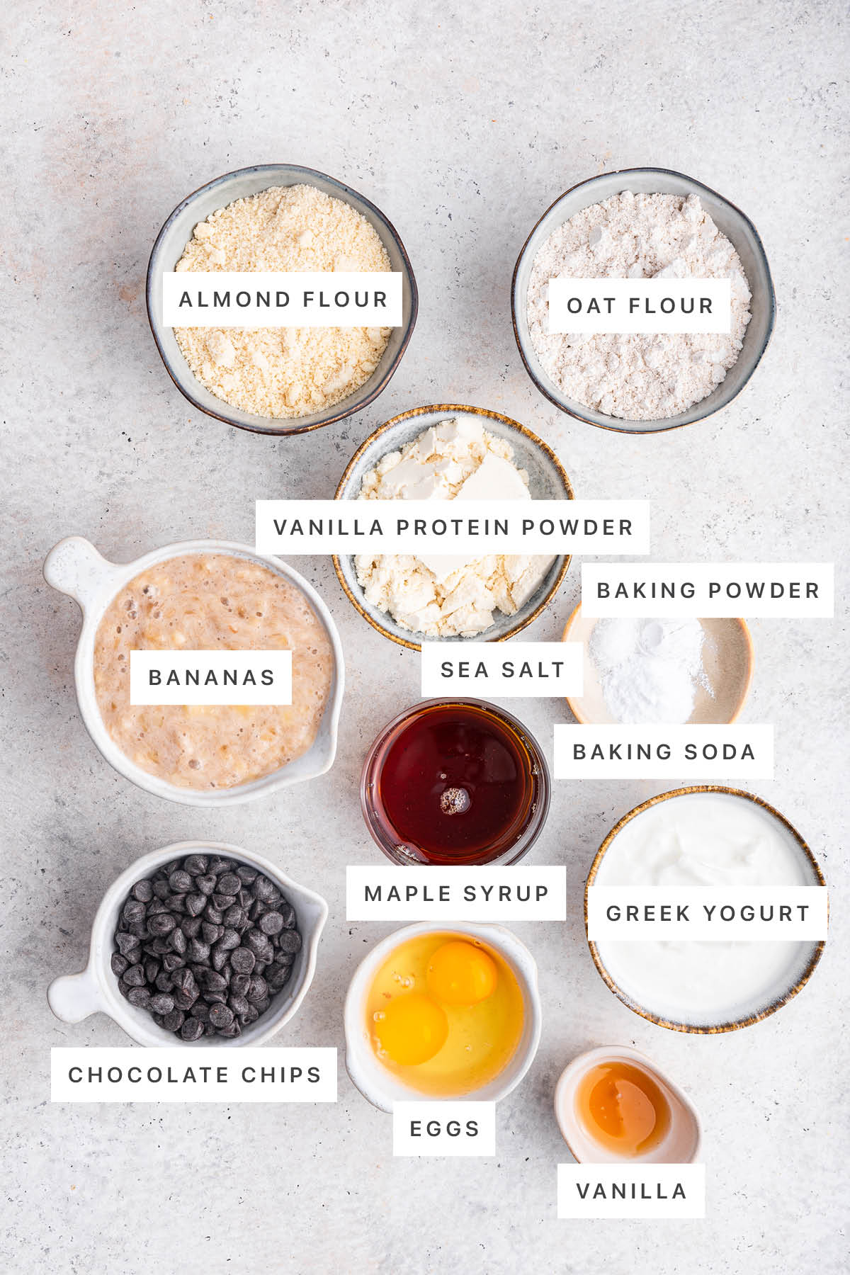 Ingredients measured out to make Protein Banana Bread: almond flour, oat flour, vanilla protein powder, baking powder, sea salt, baking soda, bananas, maple syrup, Greek yogurt, chocolate chips, eggs and vanilla.