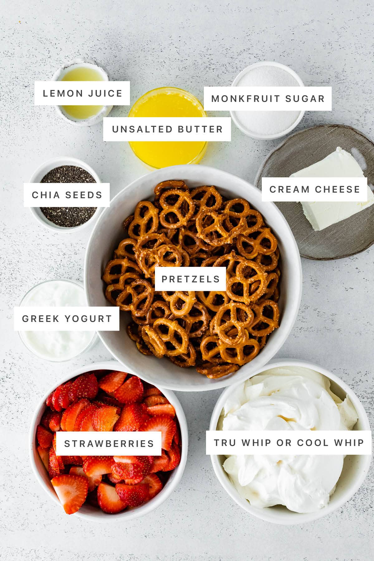 Ingredients measured out to make Healthy Strawberry Pretzel Salad: lemon juice, unsalted butter, monkfruit sugar, chia seeds, cream cheese, pretzels, Greek yogurt, strawberries and whipped topping.