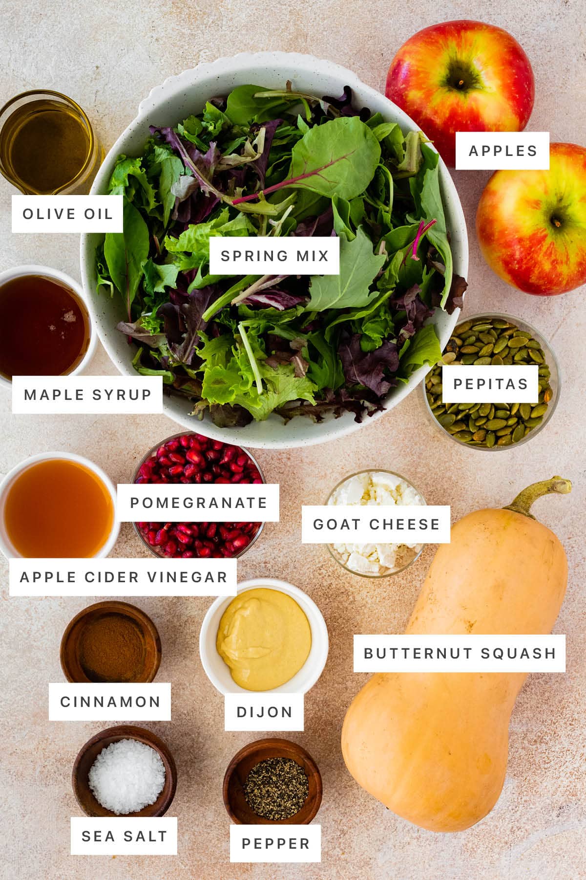 Ingredients measured out to make Thanksgiving Salad: olive oil, spring mix, apples, maple syrup, pepitas, pomegranate, goat cheese, apple cider vinegar, cinnamon, dijon, butternut squash, sea salt and pepper.