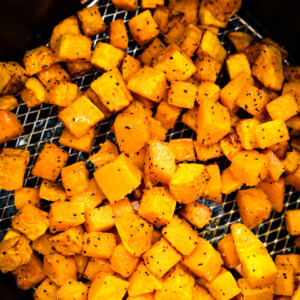 Cubed butternut squash that has been seasoned and is sitting in an air fryer basket in an air fryer.