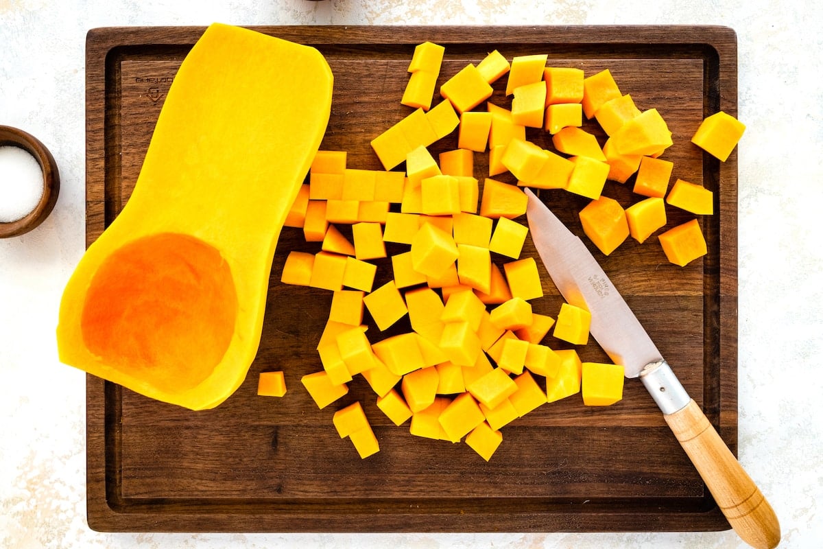 A deseeded butternut squash that has been cut in half on a wooden cutting board. One-half of the butternut squash has been cut into small cubes.