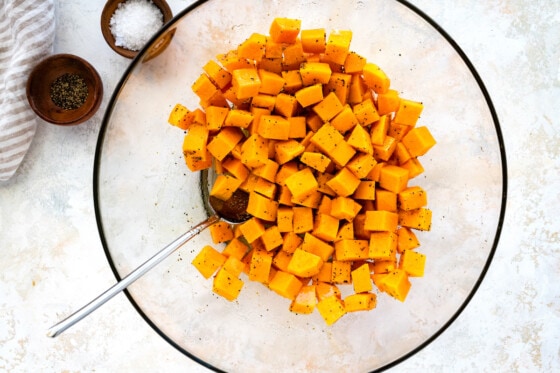 Cubed butternut squash in a large glass bowl with a metal spoon and seasoning added to the squash.