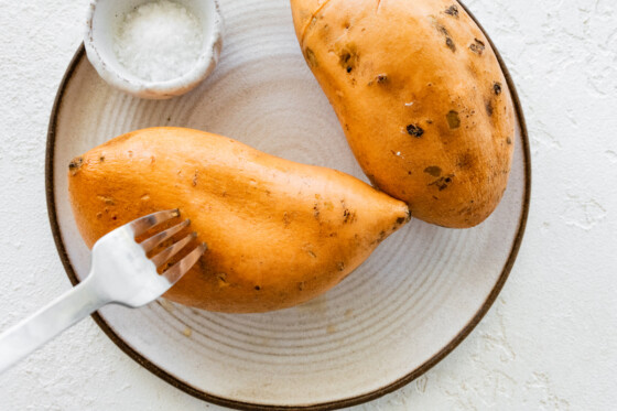 A metal fork puncturing small holes in a large sweet potato on a plate.