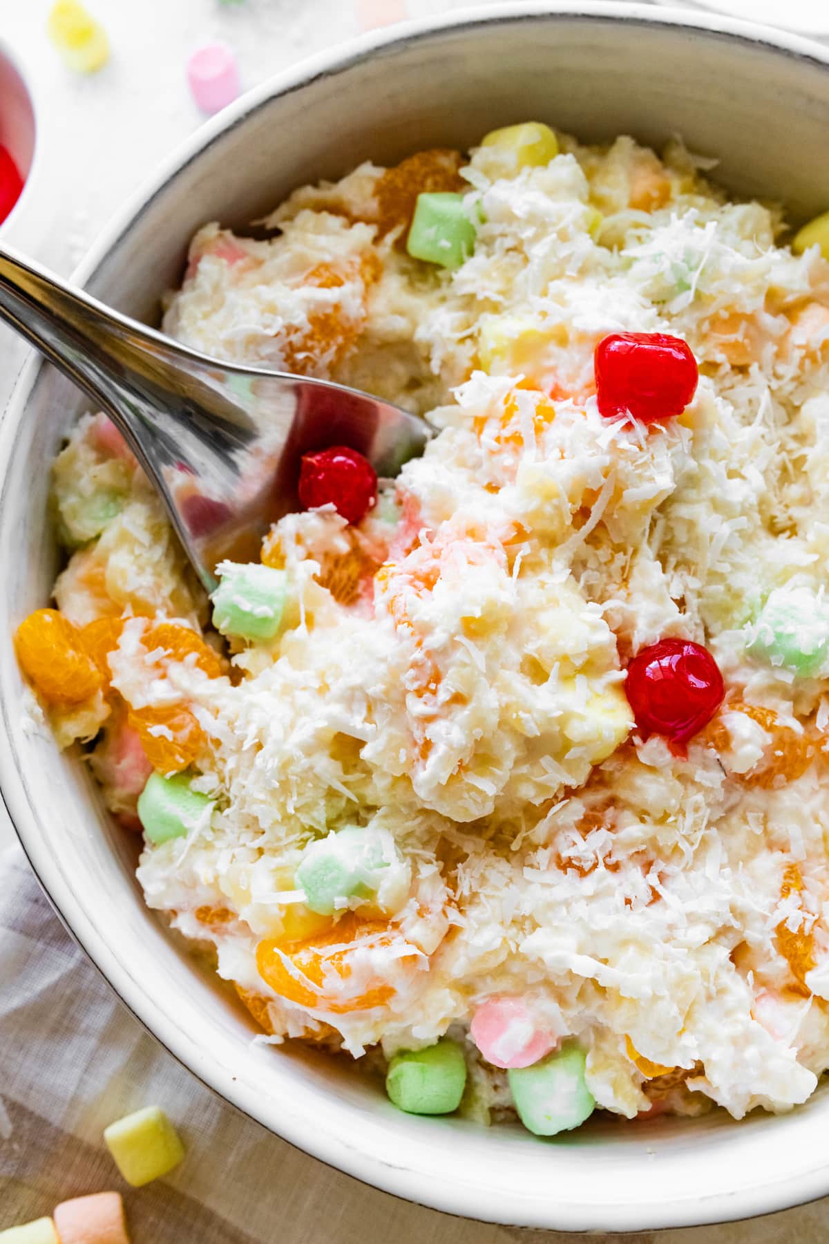 Ambrosia salad in a large bowl with a metal spoon topped with shredded coconut, cherries, and mini marshmallows.