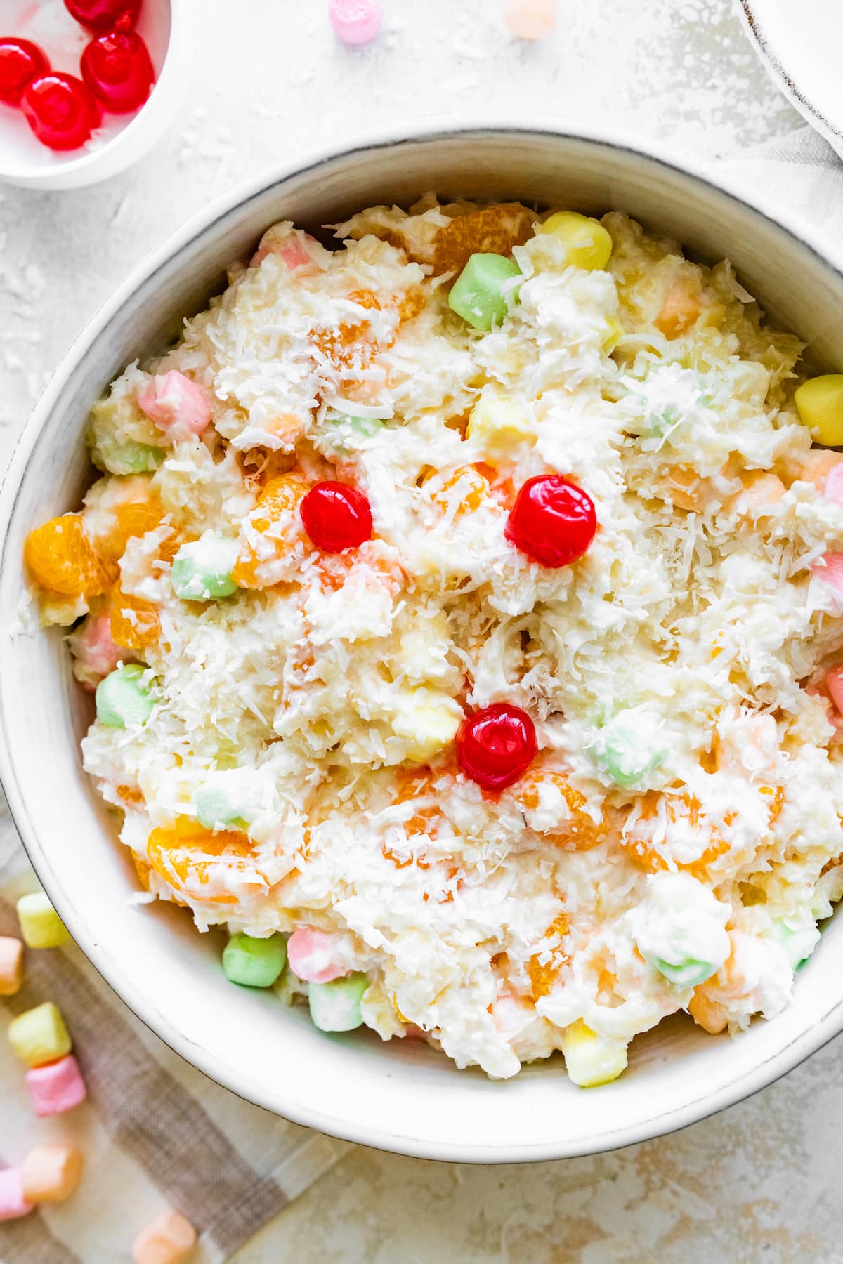 Ambrosia salad in a large bowl topped with shredded coconut, cherries, and mini marshmallows.