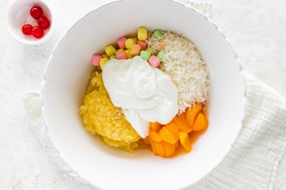 All the ingredients for an ambrosia salad are separated in a large mixing bowl. Ingredients include shredded coconut, mini marshmallows, crushed pineapple, mandarine oranges, and yogurt, with a small bowl of maraschino cherries nearby.