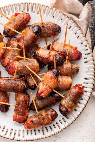 Bacon wrapped dates with toothpicks going through them on a plate.