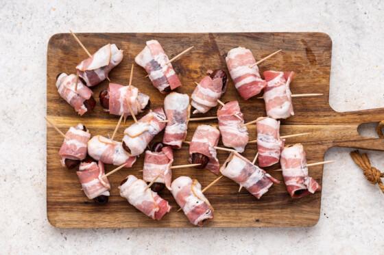 Multiple dates wrapped in raw bacon with toothpicks going through them on a wooden cutting board.