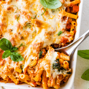 Baked ziti in a large square baking dish with a metal serving spoon taking a serving portion from the dish.