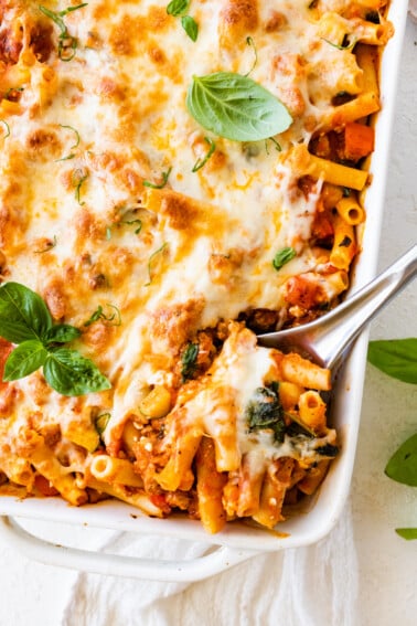 Baked ziti in a large square baking dish with a metal serving spoon taking a serving portion from the dish.