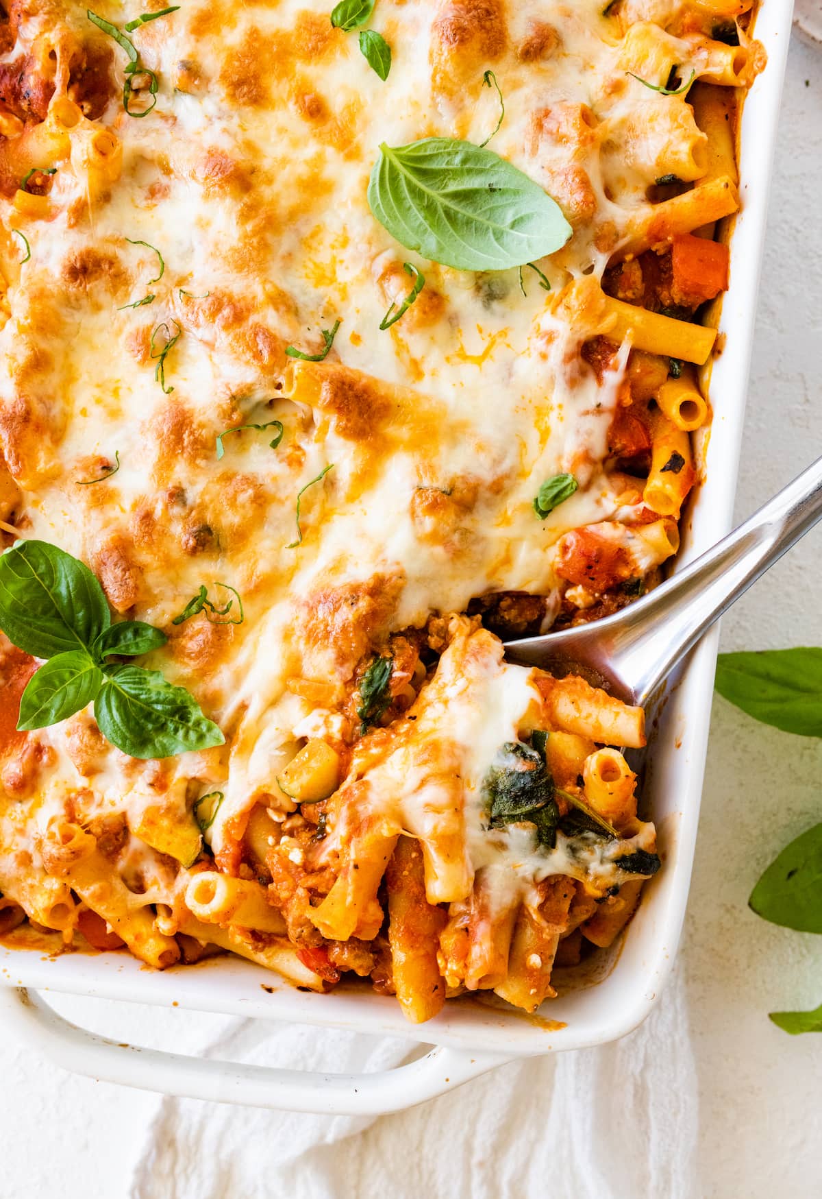 Baked ziti in a large square baking dish with a metal serving spoon taking a serving portion from the dish.