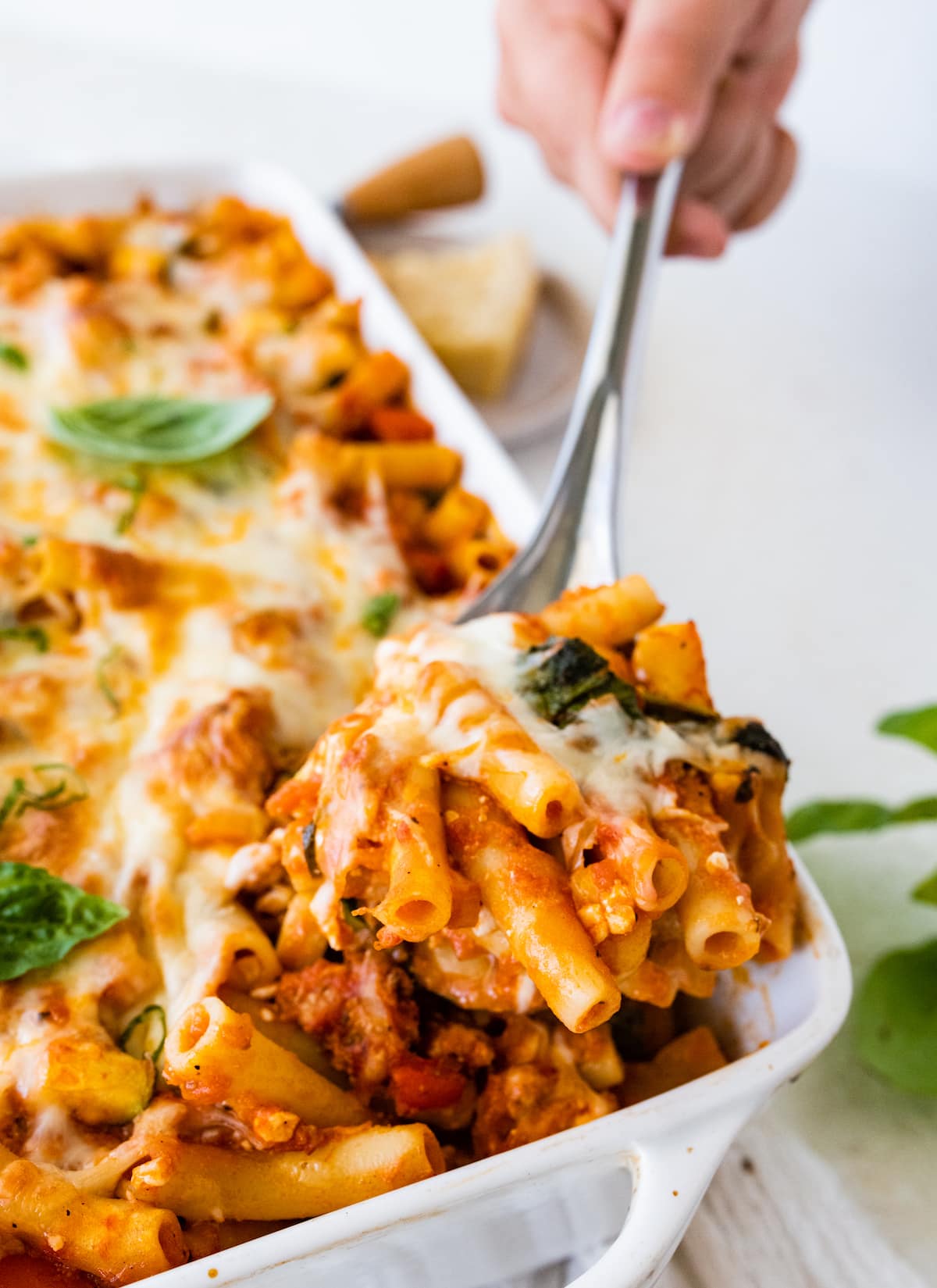 A hand uses a metal serving spoon to take a serving size of baked ziti from a square baking dish.