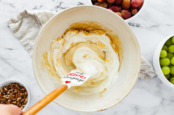 A woman's hand uses a silicon spatula to mix cream cheese, sour cream, sugar, and vanilla extract that will be used for a grape salad in a large mixing bowl.
