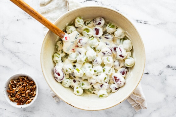Red and green grapes covered in the yogurt and sour cream mixture in a large bowl with a silicone spatula. Near the bowl is a small bowl of crushed pecans.