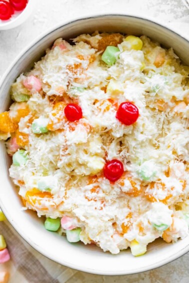 Ambrosia salad in a large bowl topped with shredded coconut, cherries, and mini marshmallows.
