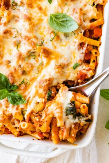 Baked ziti in a large square baking dish with a metal serving spoon taking a serving portion from the dish.
