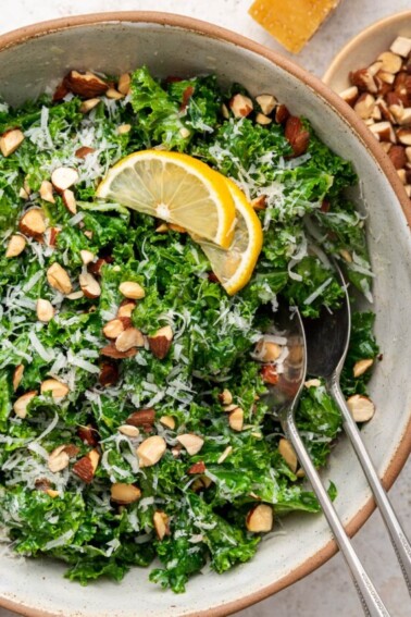 Kale salad in a serving bowl with serving utensils topped with almonds, parmesan cheese and lemon slices.