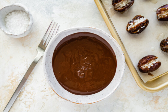 A small bowl of melted chocolate near a baking tray with Medjool dates that have been filled with coconut butter and coconut shreds.