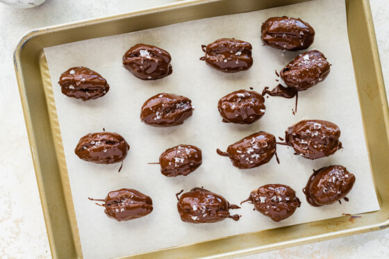 Multiple dates covered in melted chocolate and a sprinkle of flaky sea salt on a baking tray.