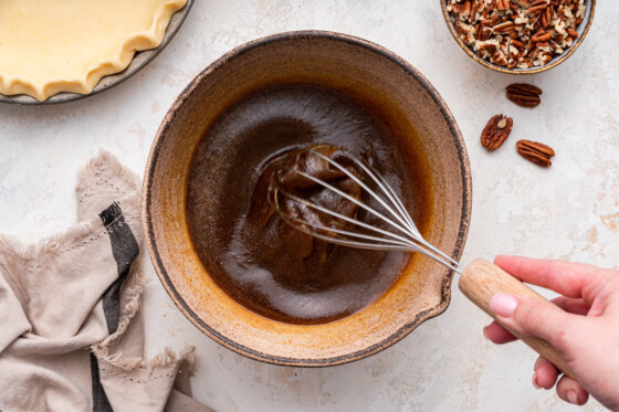 A woman's hand whisking the remaining wet ingredients for the pecan pie in a mixing bowl.