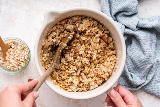 A woman's hand uses a large wooden spoon to combine ingredients for granola in a large mixing bowl.