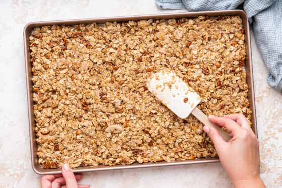 A woman's hand uses a silicone spatula to spread granola ingredients on a baking sheet.
