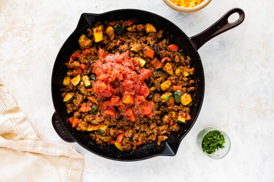 Roasted tomatoes added to a large cast iron skillet with ground beef, zucchini, red bell pepper, garlic, and onion.