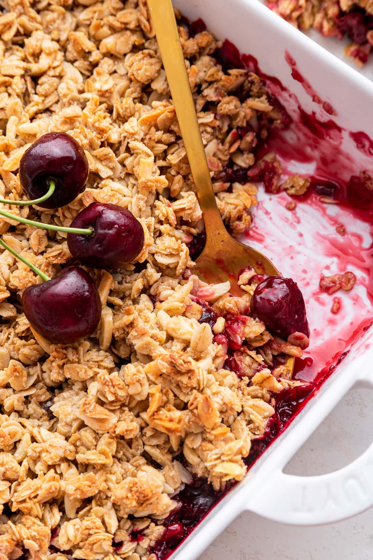 Cherry crisp in a square baking dish with whole cherries on top and a metal spoon taking a small serving.