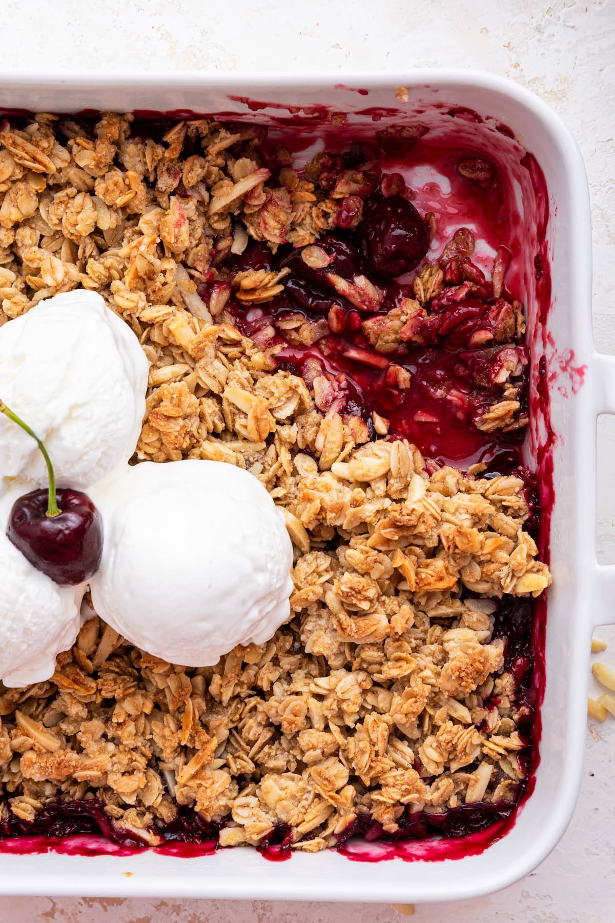 Cherry crisp in a square baking dish with three scoops of vanilla ice cream and a cherry in the middle. There is a serving of cherry crisp missing from a corner of the baking dish.