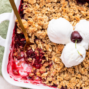 Cherry crisp in a square baking dish with three scoops of vanilla ice cream and a whole cherry on top in the middle of the crisp. There's also a metal serving spoon in the corner of the baking dish where a serving of the cherry crisp has been taken.