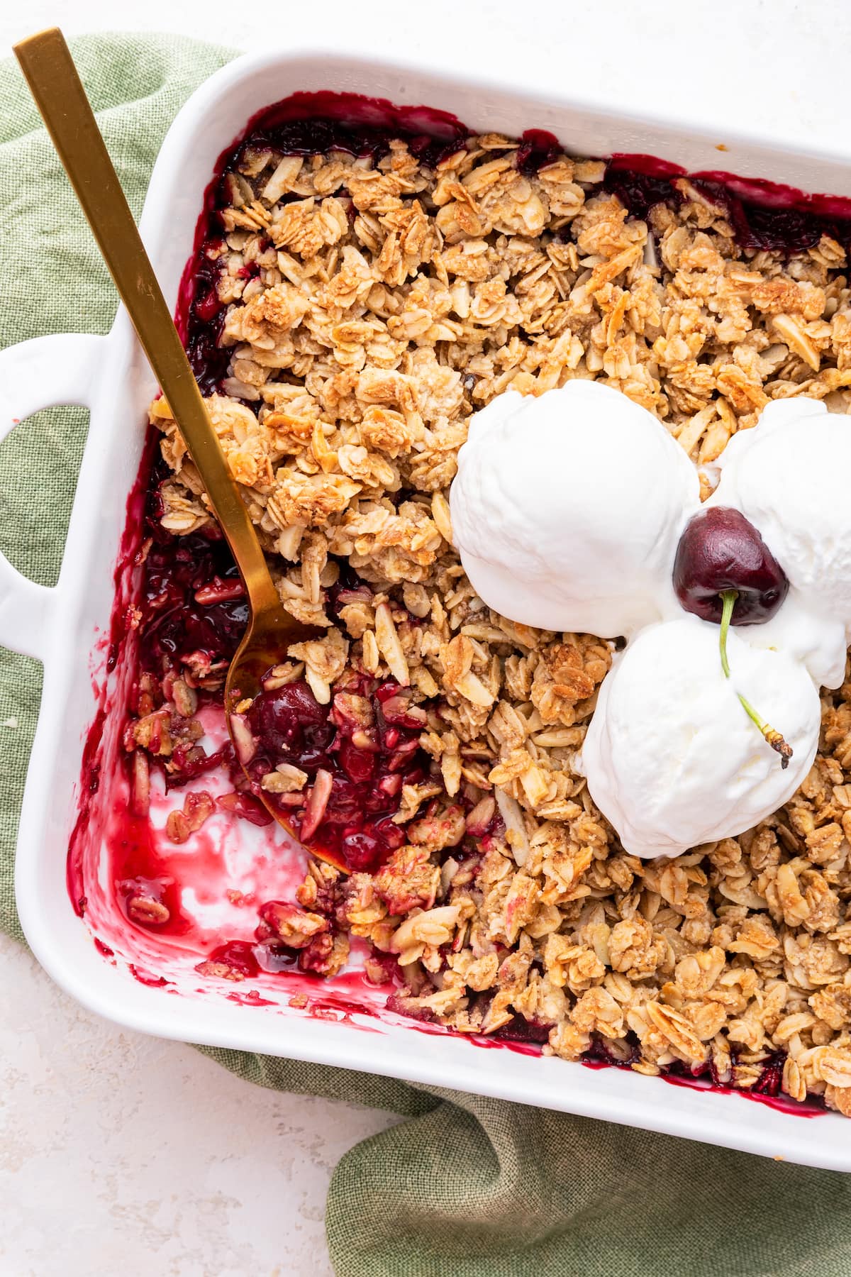 Cherry crisp in a square baking dish with three scoops of vanilla ice cream and a whole cherry on top in the middle of the crisp. There's also a metal serving spoon in the corner of the baking dish where a serving of the cherry crisp has been taken.