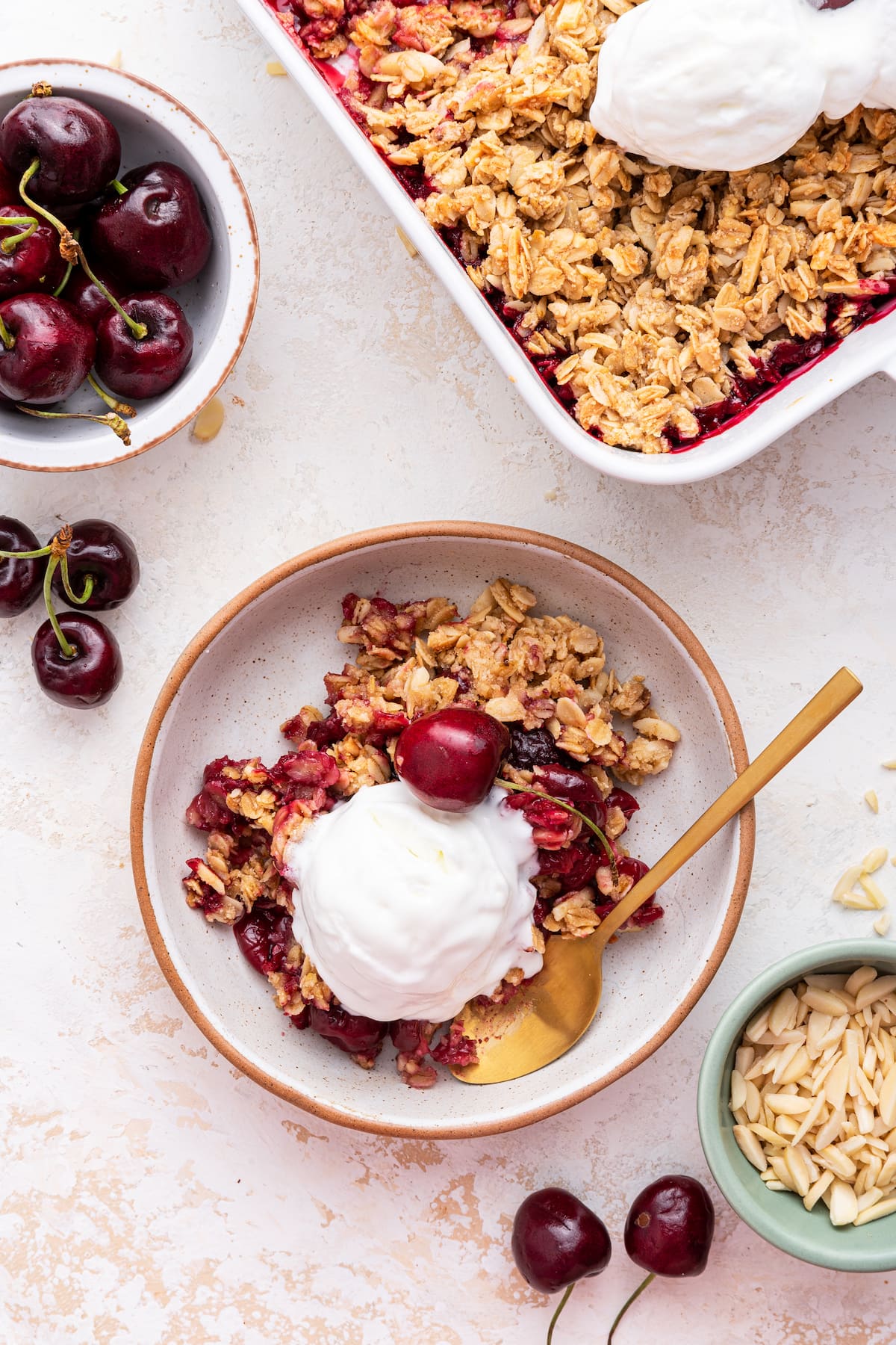 A serving of cherry crisp on a small plate with a metal spoon, a scoop of vanilla ice cream, and a cherry on top.