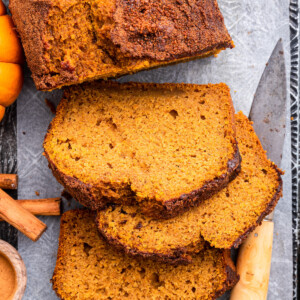 A loaf of healthy pumpkin bread on parchment paper on a cutting board. There's three slices of bread near the loaf with a cutting knife, three cinnamon sticks, and small pumpkins also nearby.