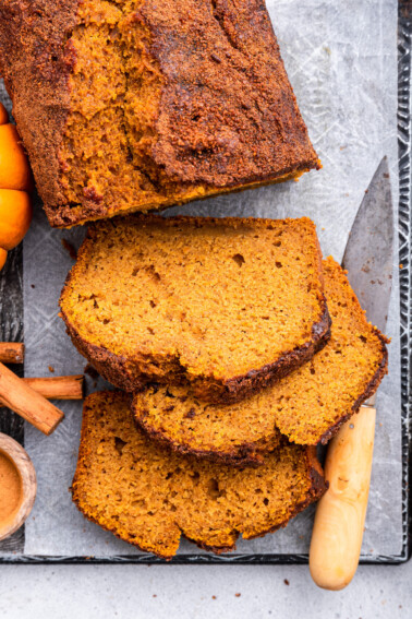 A loaf of healthy pumpkin bread on parchment paper on a cutting board. There's three slices of bread near the loaf with a cutting knife, three cinnamon sticks, and small pumpkins also nearby.