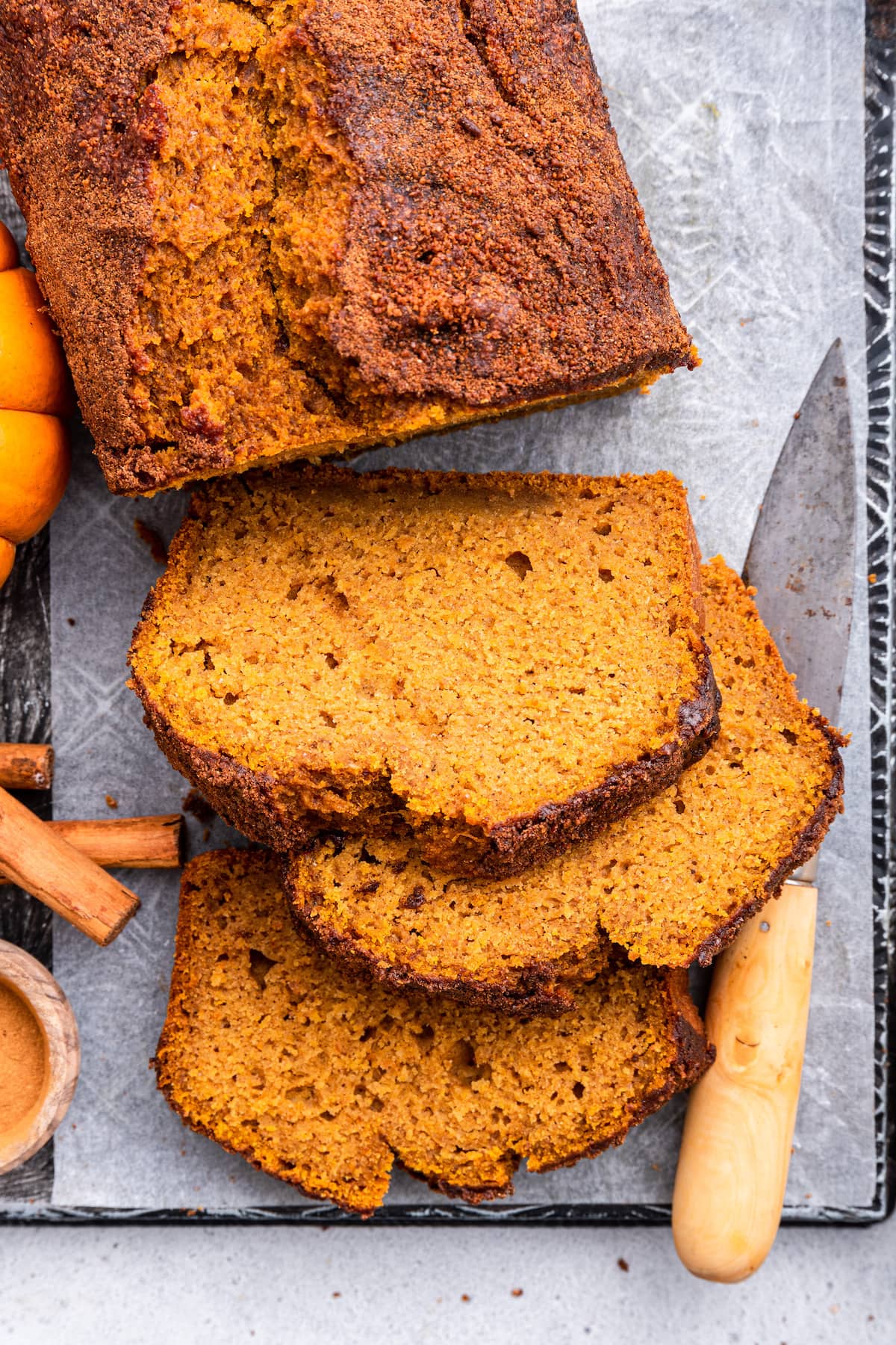 A loaf of healthy pumpkin bread on parchment paper on a cutting board. There's three slices of bread near the loaf with a cutting knife, three cinnamon sticks, and small pumpkins also nearby.