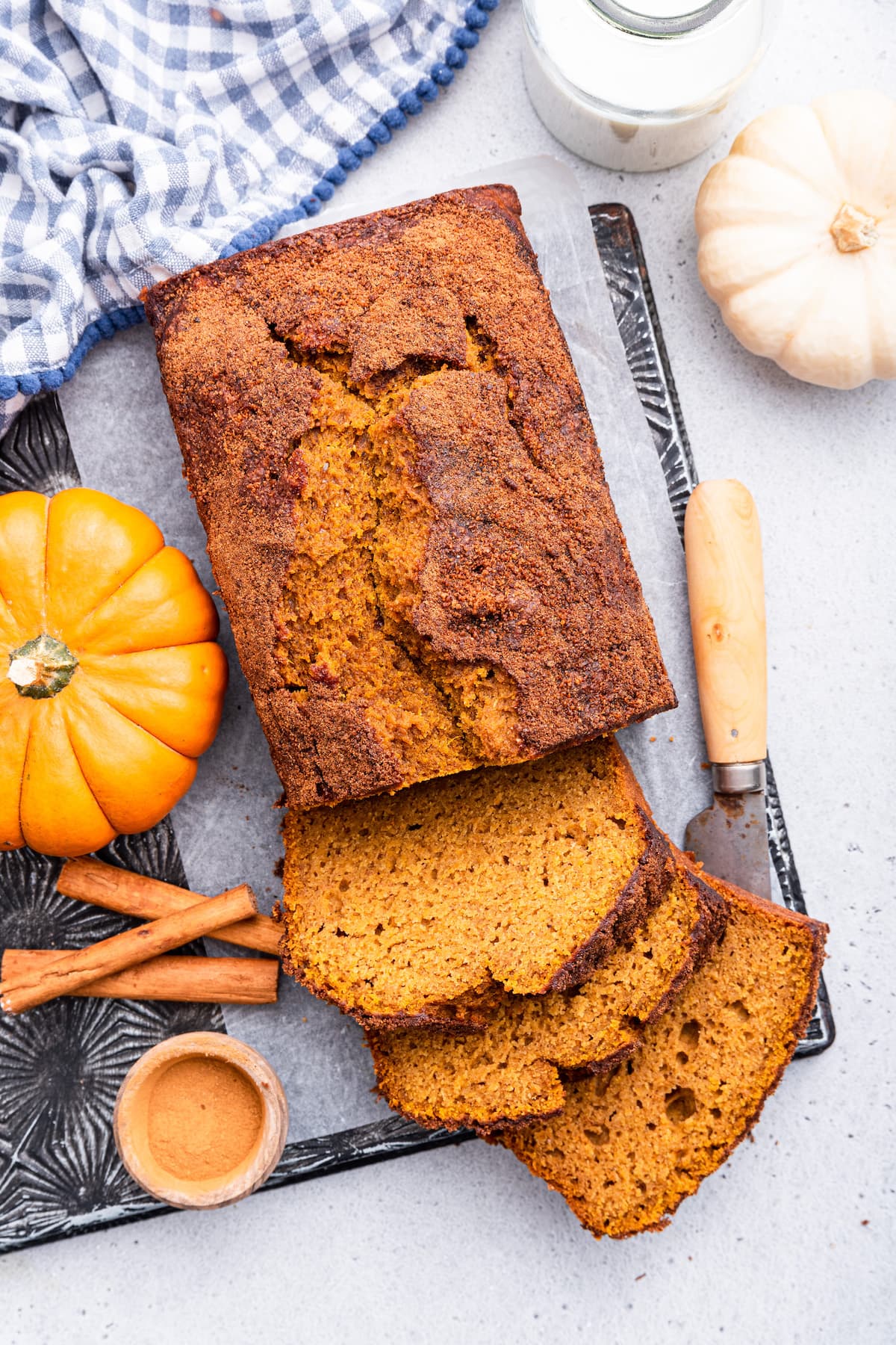 A loaf of healthy pumpkin bread on parchment paper on a cutting board. There's three slices of bread near the loaf with a cutting knife, three cinnamon sticks, and small pumpkins also nearby.