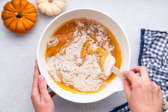 A woman's hand uses a silicone spatula to combine the wet and dry ingredients for the healthy pumpkin bread in a large mixing bowl.