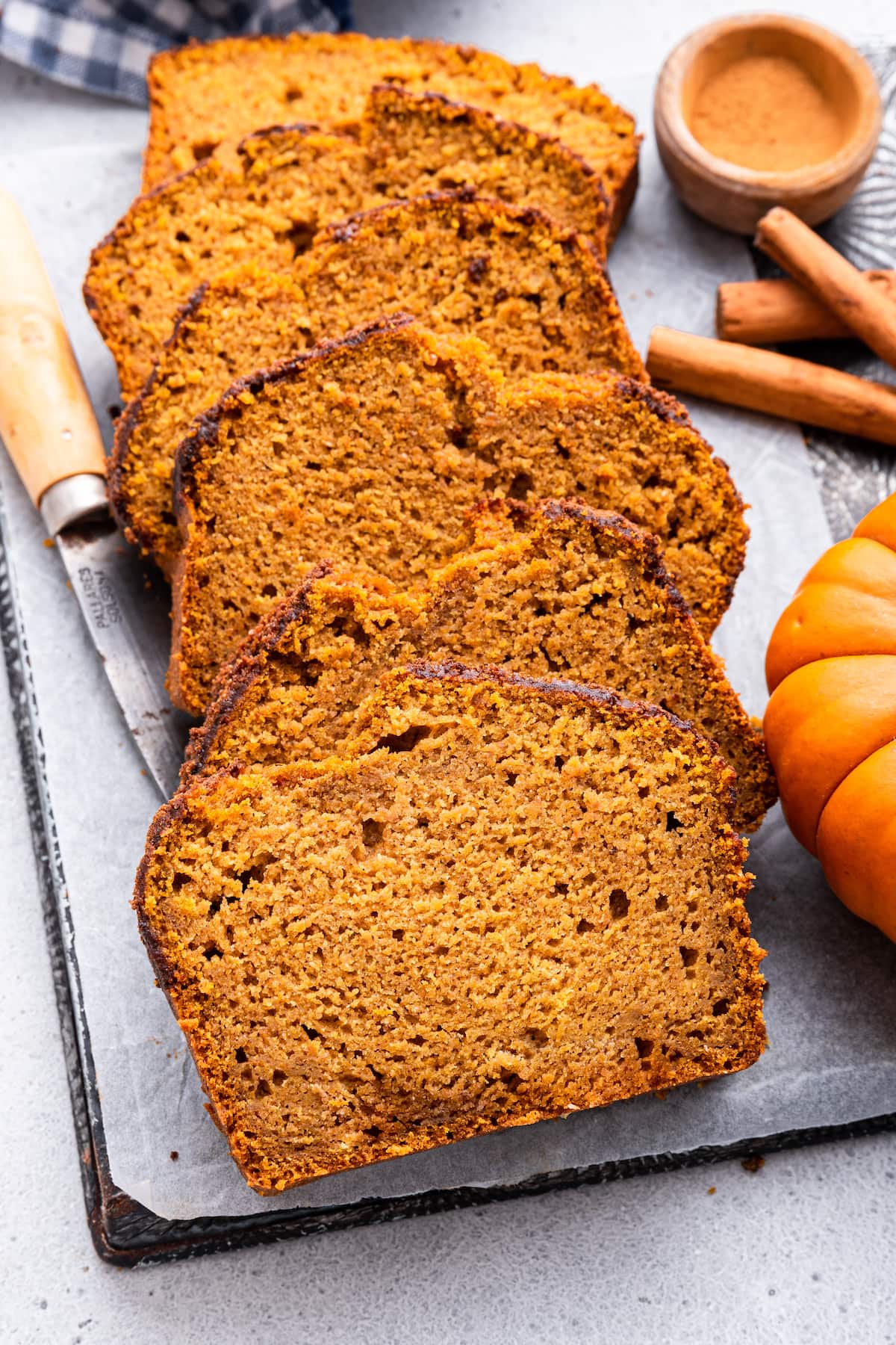 Multiple slices of healthy pumpkin bread leaning against one another on a cutting board with a knife, cinnamon sticks, and a small pumpkin nearby.