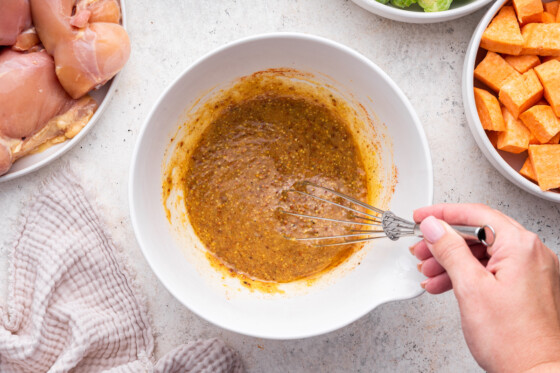 A woman's hand using a metal whisk to whisk the honey mustard marinade in a mixing bowl for the chicken.