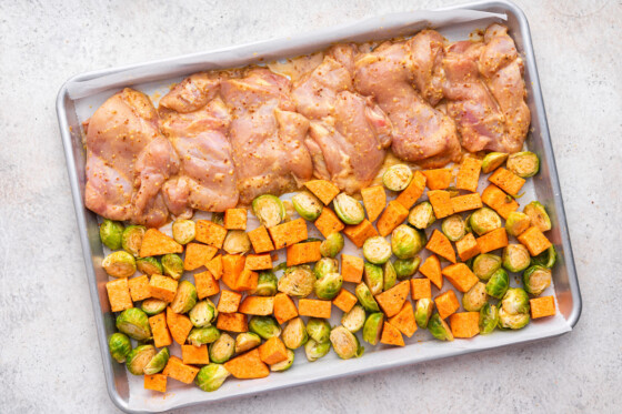 Multiple chicken thighs covered in a honey mustard marinade on one side of a large sheet pan, with the other side containing brussels sprouts and diced sweet potato.