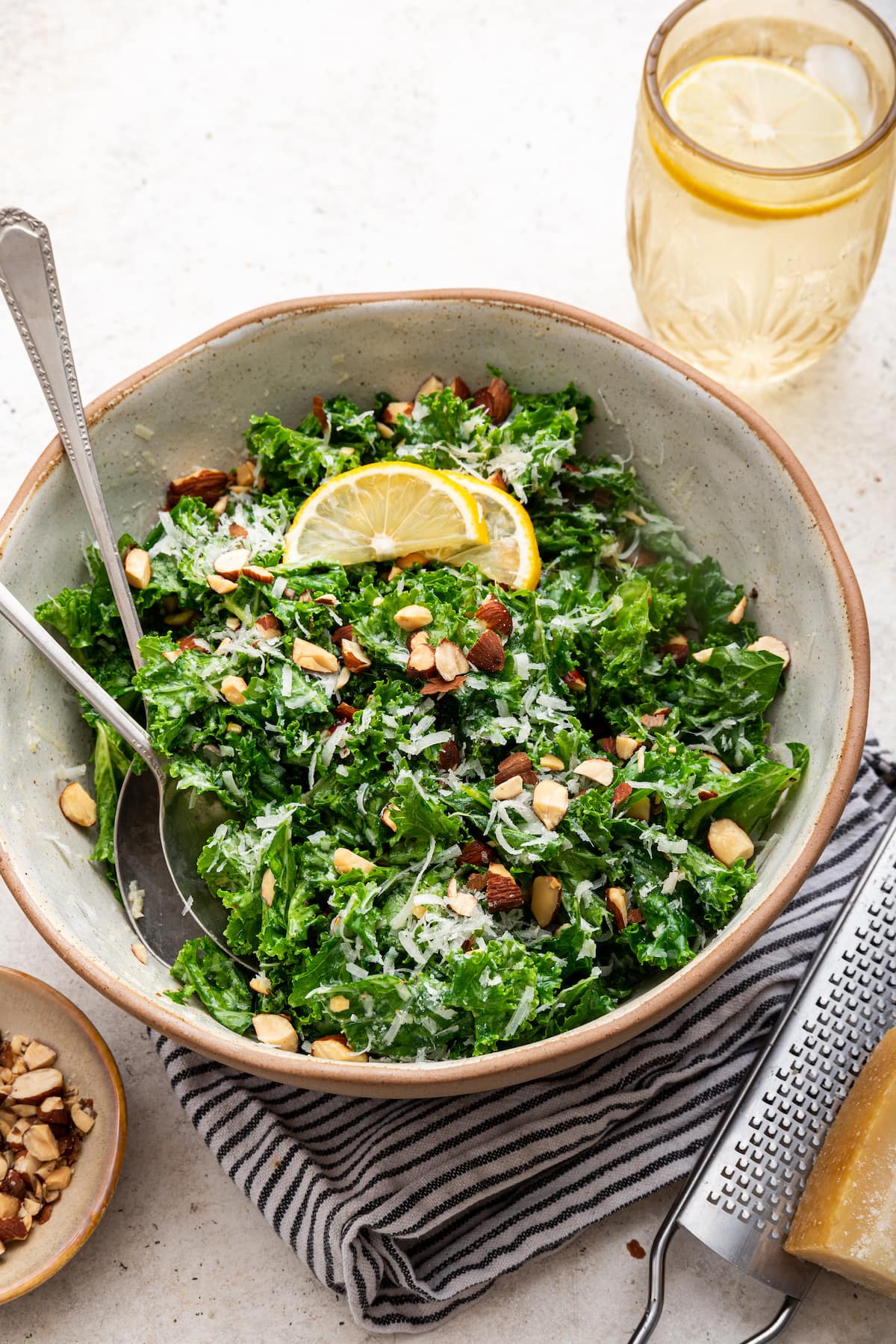 Kale salad in a serving bowl with serving utensils topped with almonds, parmesan cheese and lemon slices.