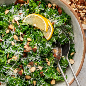 Kale salad in a serving bowl with serving utensils topped with almonds, parmesan cheese and lemon slices.