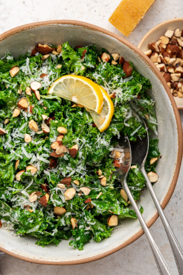 Kale salad in a serving bowl with serving utensils topped with almonds, parmesan cheese and lemon slices.