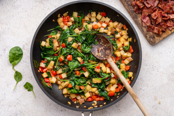 Diced bell pepper, potatoes, and wilted spinach in a large skillet with a wooden spoon.