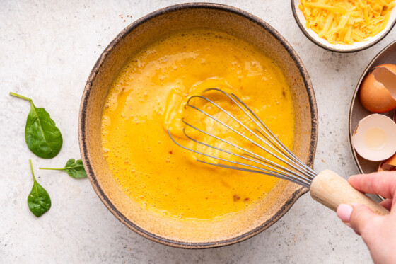 A woman's hand uses a metal whisk to whisk eggs in a large mixing bowl.