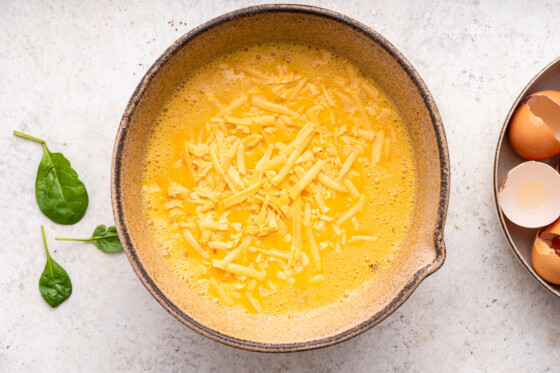 Shredded cheddar cheese added to a large mixing bowl of eggs.