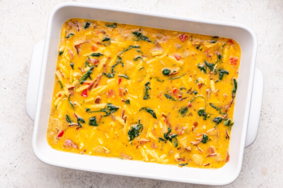 Eggs are added to a large baking dish, vegetables, and turkey bacon to make a casserole.