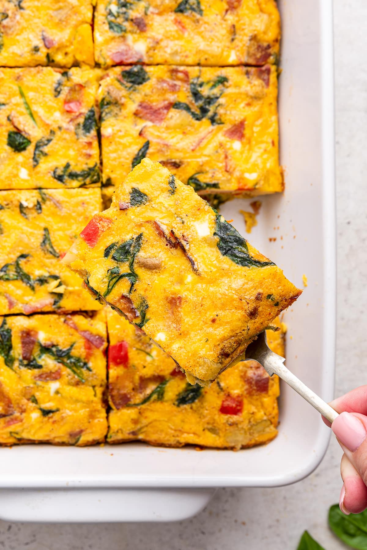 A woman's hand uses a metal spatula to lift a piece of egg casserole away from a square baking dish with the rest of the casserole in it.