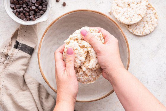 Hands are holding a ball of crushed rice cakes over a large mixing bowl used for no bake peanut butter crunch bars.